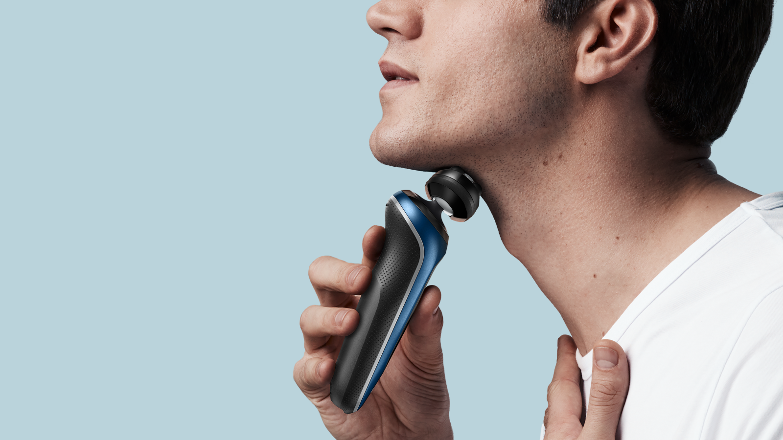 Half of a profile shot of a man in white shirt shaving his face, with shaver device in left hand, his right hand is placed under the neck area for support