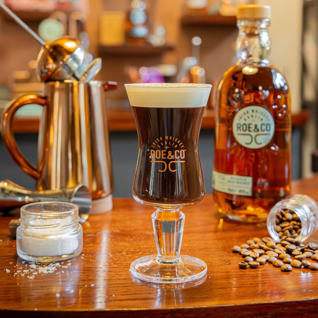 Roe & Co Irish whiskey coffee cocktail in branded glass, with bottle, coffee beans, sugar and bar tools on wooden bar.