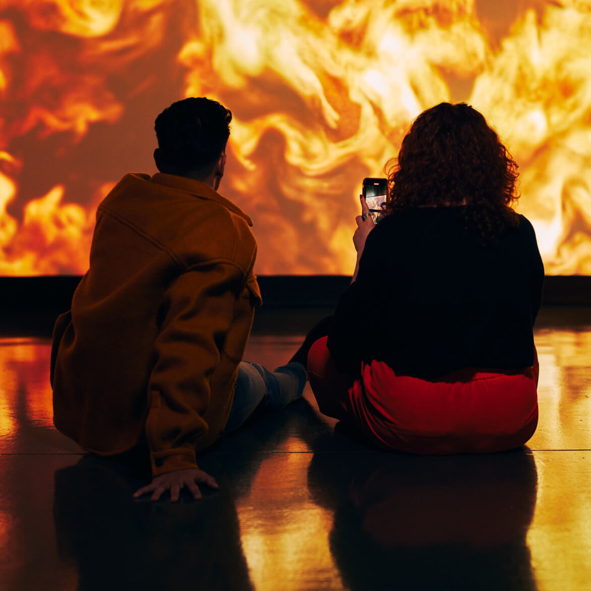 Two people sitting on the ground of the cinema room within the Guinness Storehouse, the girl on the right is recording the images on the screen.,