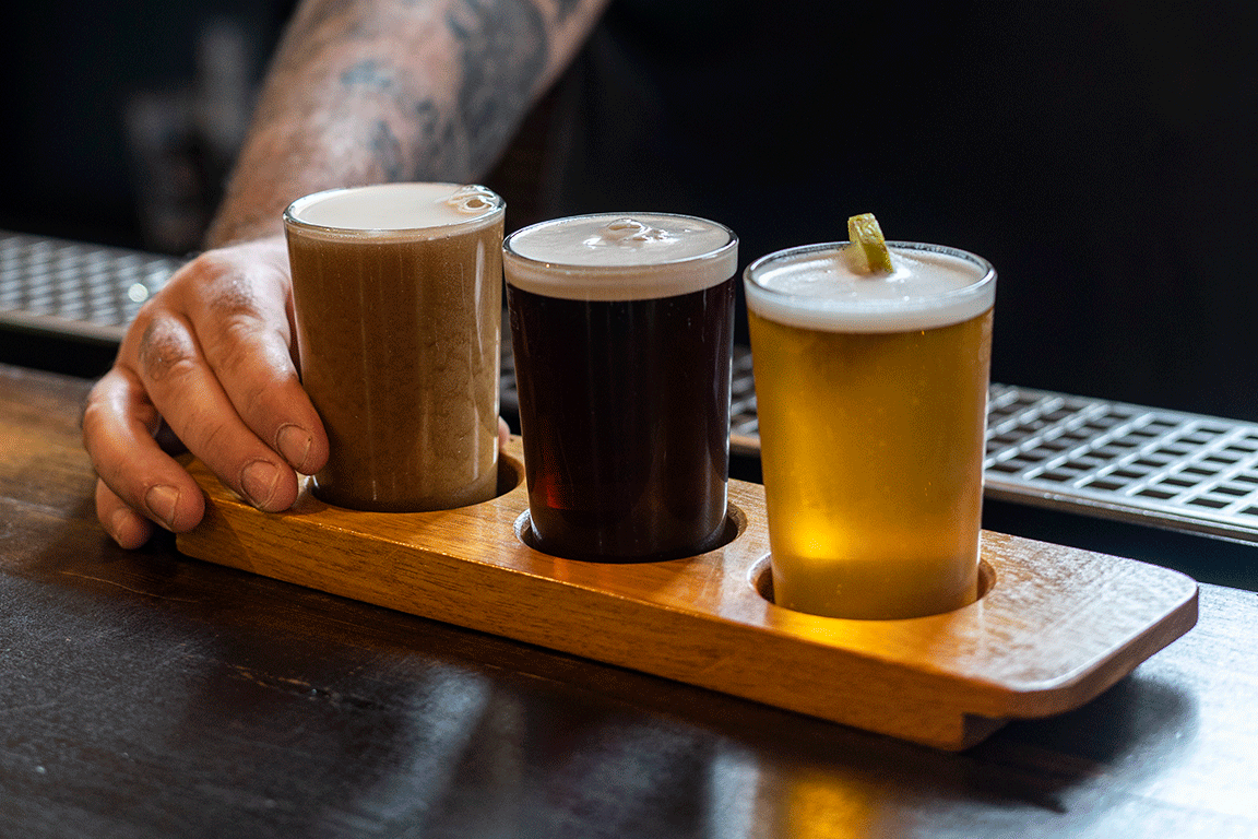 A paddle of three beers of various colours on a bar counter, with a hand behind them.