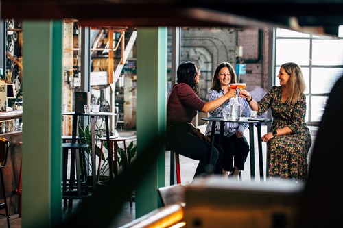 Three ladies in a bar sitting on a high stool cheering with their lovely cocktails