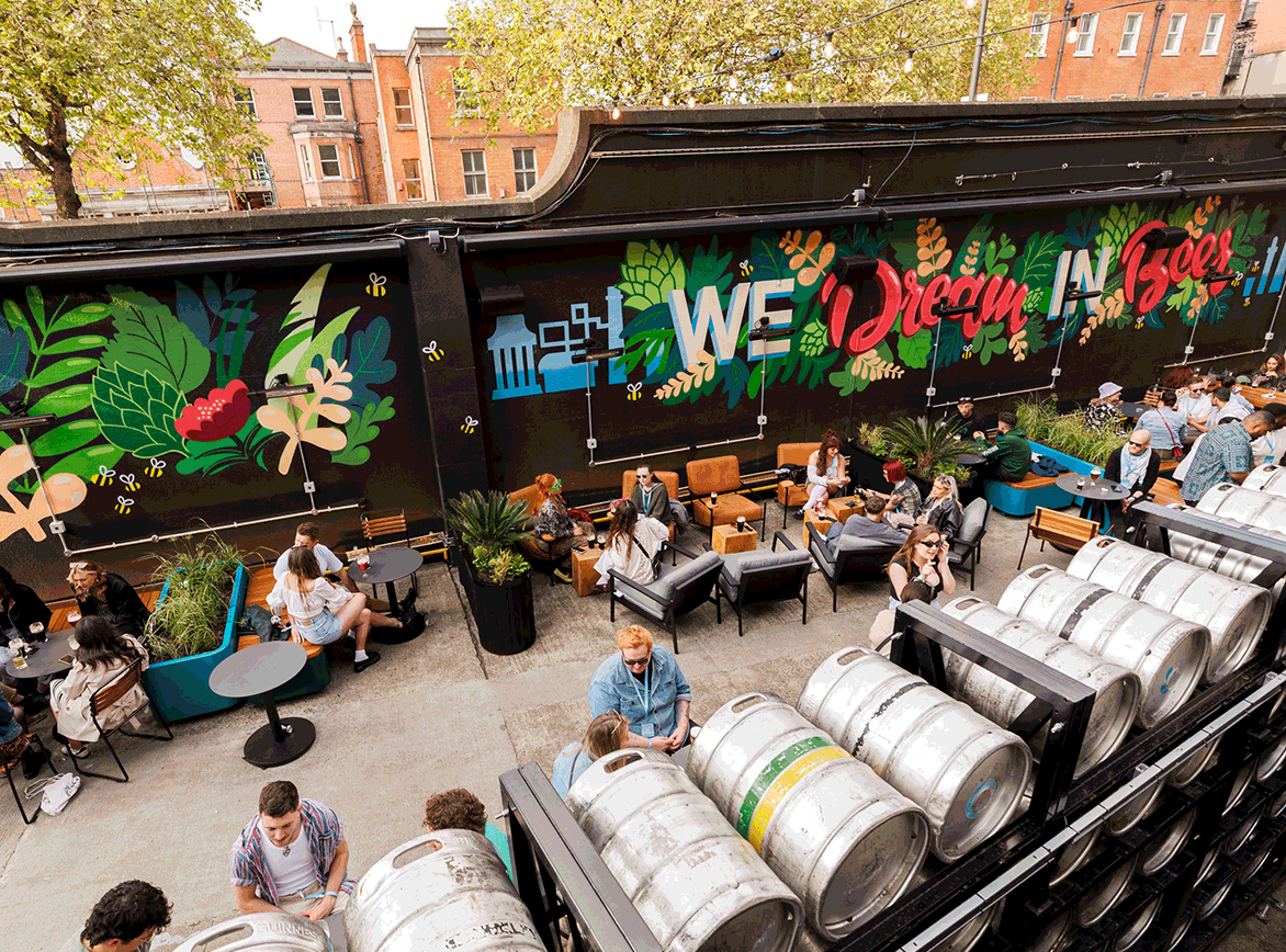 The outside area at Guinness Open Gate Brewery, with people at tables drinking beers and water, with huge kegs in the foreground