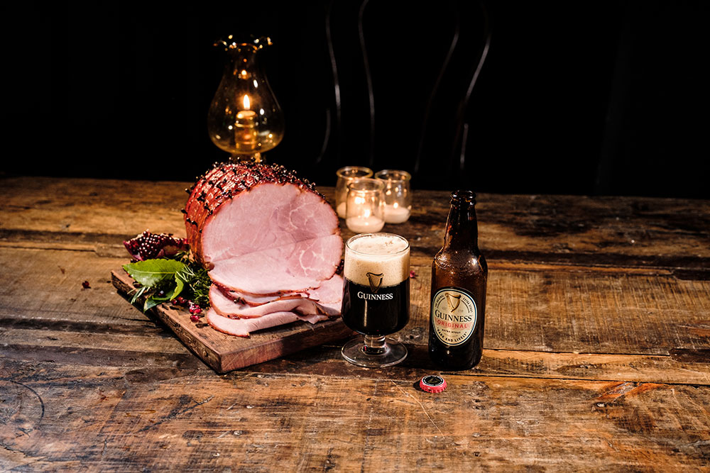 A Guinness cured ham on a wooden table with a glass of Guinness original beside it