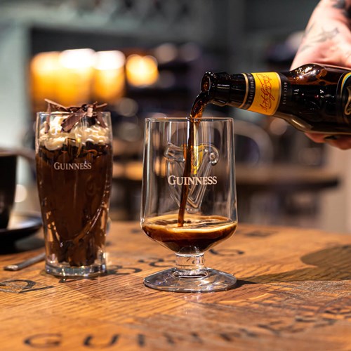 A bottle of Guinness Foreign Extra Stout being poured into a Guinness embossed stem glass with a dessert in a Guinness pint glass beside it.