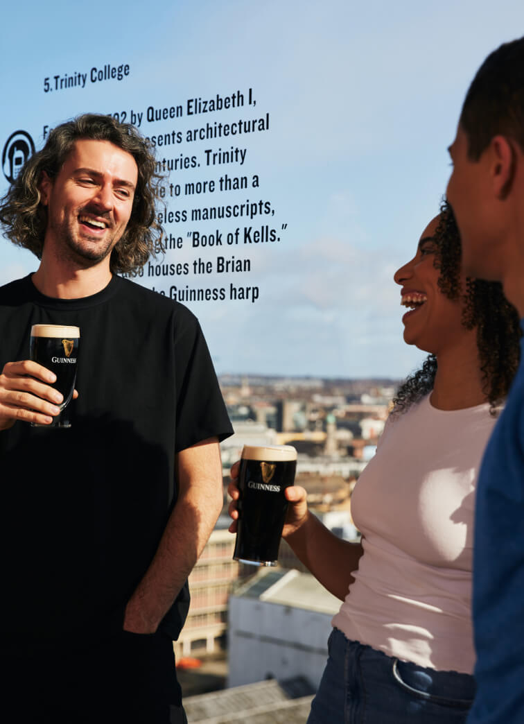 Two men and one woman smiling and chatting while holding perfectly poured pints of creamy Guinness. The background is a breath-taking view of Dublin