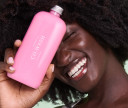 Happy woman with healthy, coily hair showing her customized Co-wash bottle in pink color.