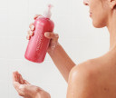 Woman holds her customized pink shampoo up in shower in right hand. Left hand has lathered shampoo in it, showing gentle foaming of shampoo. 