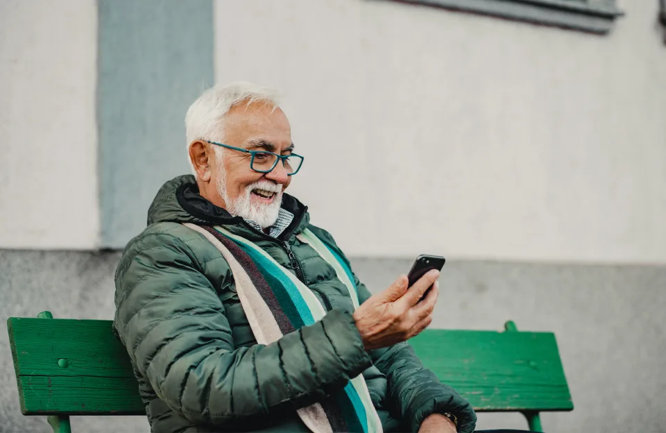 Älterer Herr sitzt auf einer grünen Bank und surft im Internet mit dem Smartphone