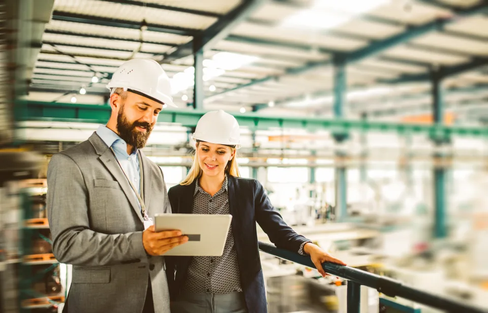 Zwei Arbeitskollegen tragen einen Helm und schauen auf ein Tablet während sie in einer Fabrik