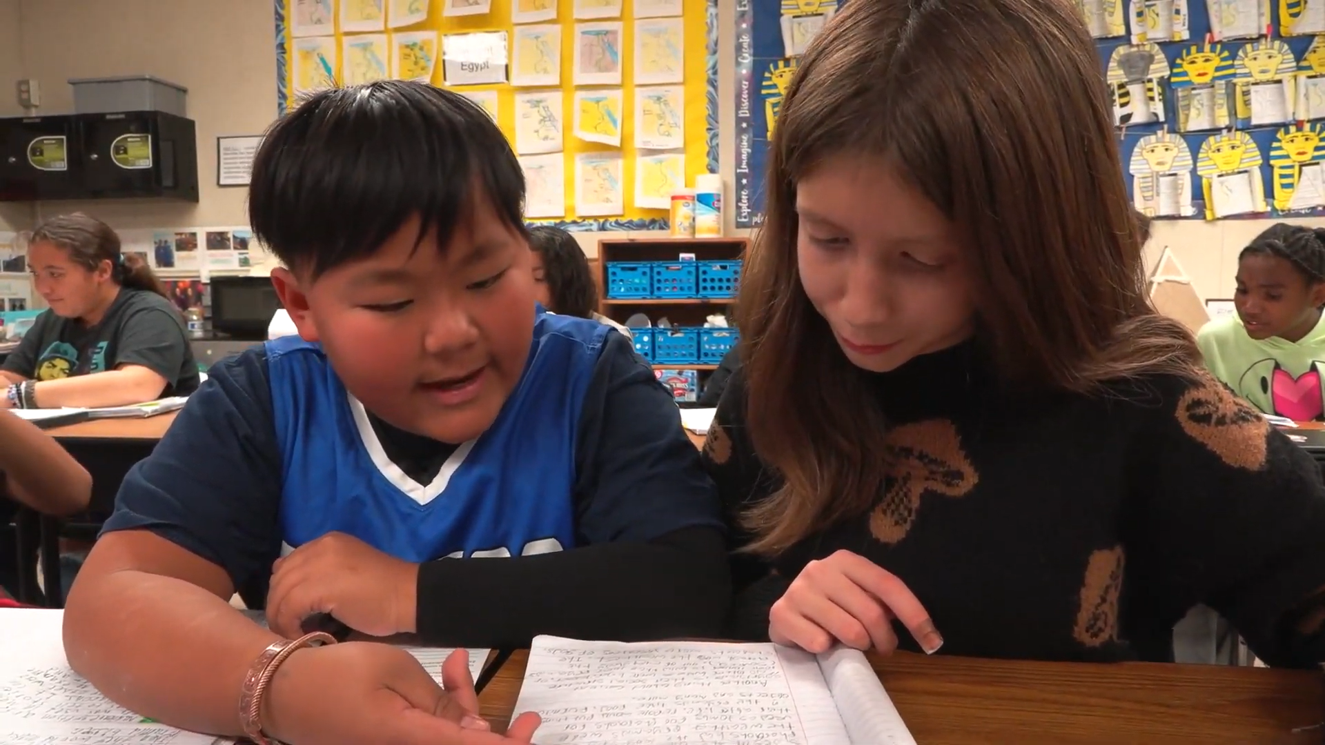 Two students sit side by side at a classroom table, looking at a shared notebook and discussing their work, with other classmates studying in the background.
