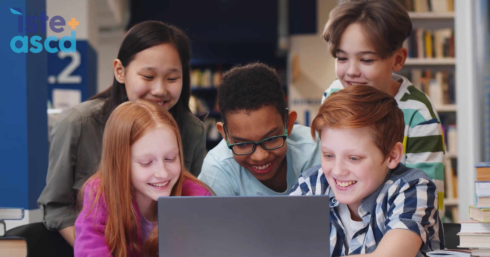 A group of smiling students gathers around a laptop in a library, engaged and collaborating on a project.