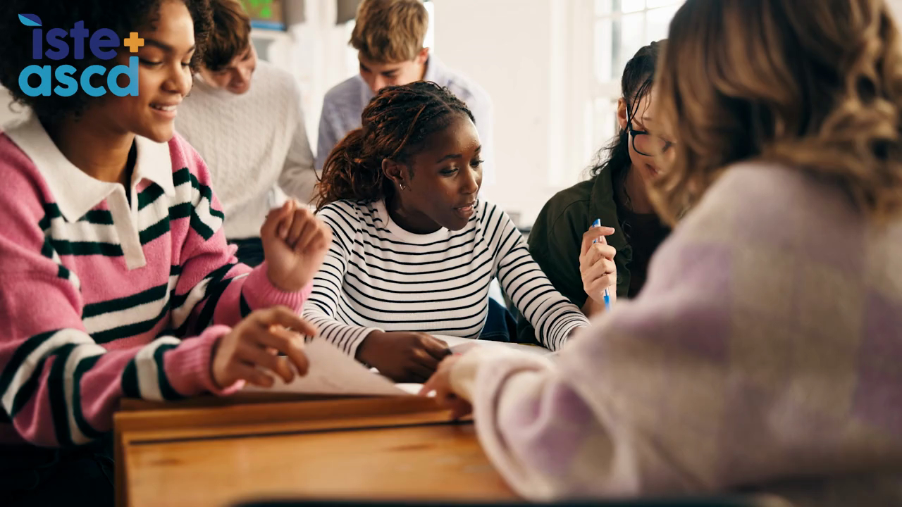 Video still of high school students sitting around a table discussing a topic together and asking questions.