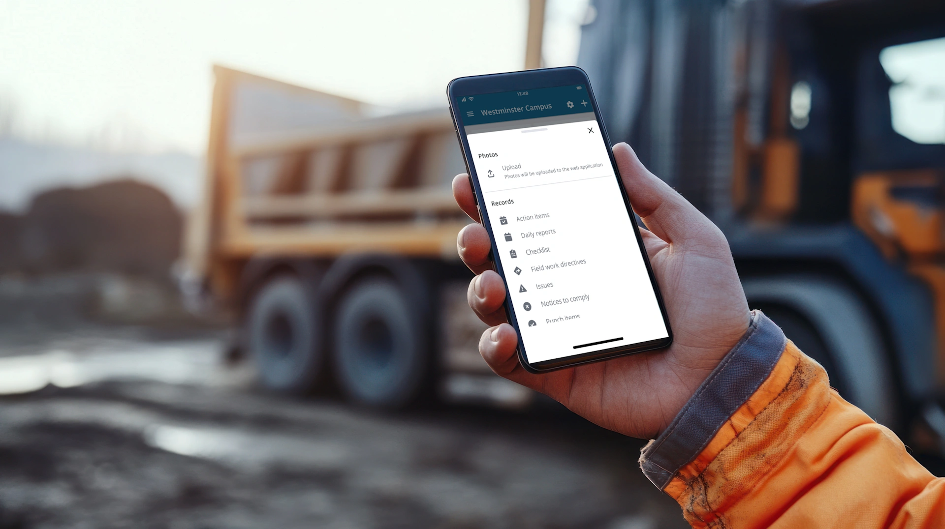 A hand in high-visibility clothing holds a smartphone displaying the Trimble ProjectSight mobile app for the Westminster Campus project at a construction site.