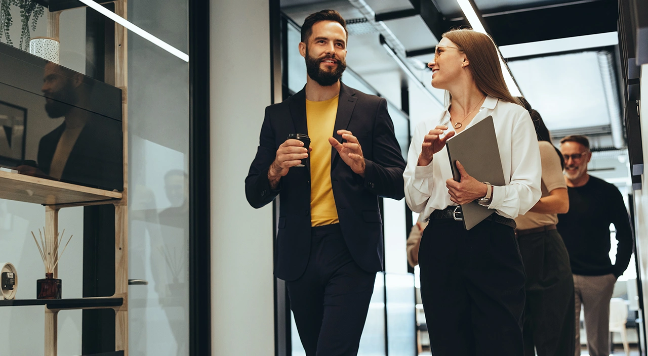 Colleagues walking and talking in a modern office hallway. A man holds a coffee cup while a woman carries a laptop.