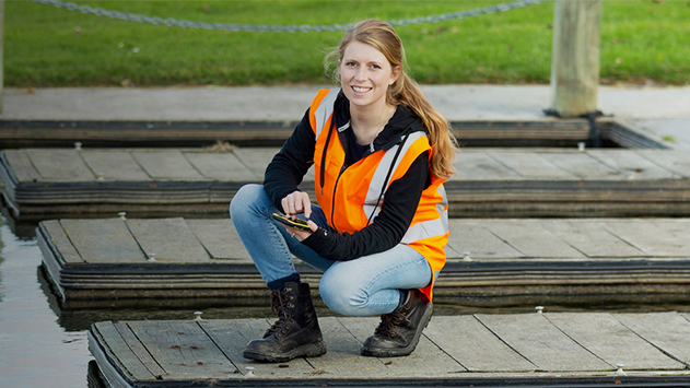 Woman in reflective vest and boots on dock smiling at camera using mobile device