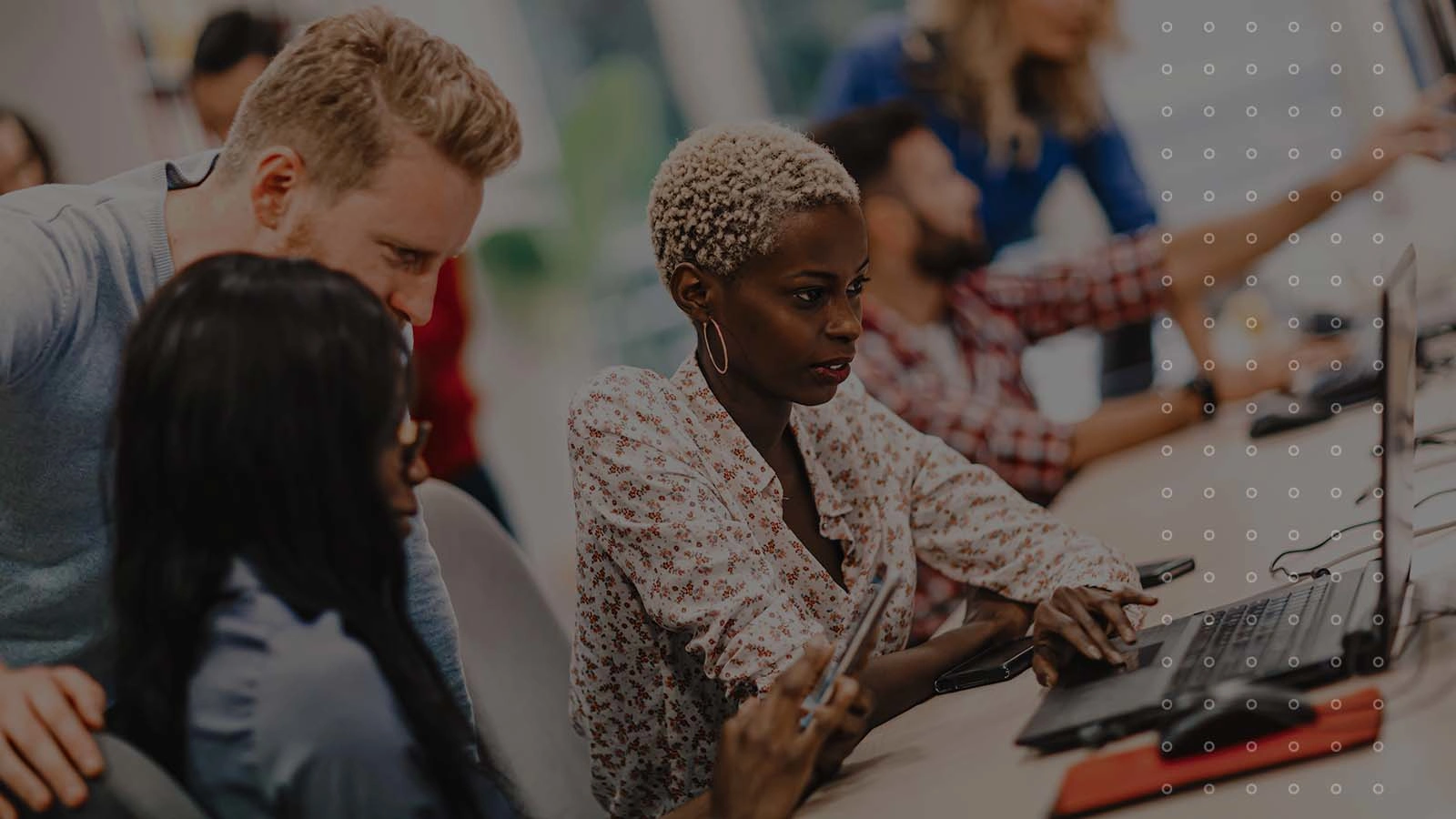 A diverse team of professionals, including a man with red hair and a woman with short blonde hair, collaborate around a laptop in an office.