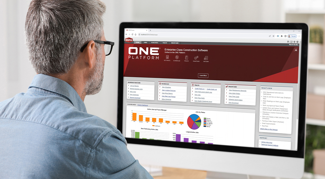 A man using Trimble's B2W ONE Platform construction software on a computer, viewing project dashboards and performance charts.