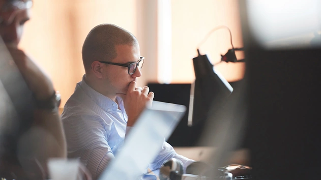 A man in a blue shirt and glasses sits at a desk, contemplating a task with one hand on his chin.