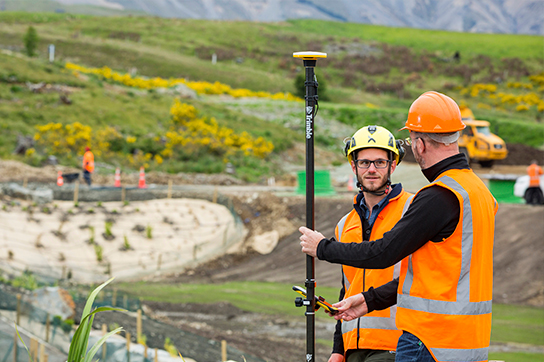 Two construction or surveying workers in hard hats and safety vests discussing plans with a Trimble GPS surveying pole on a development site.