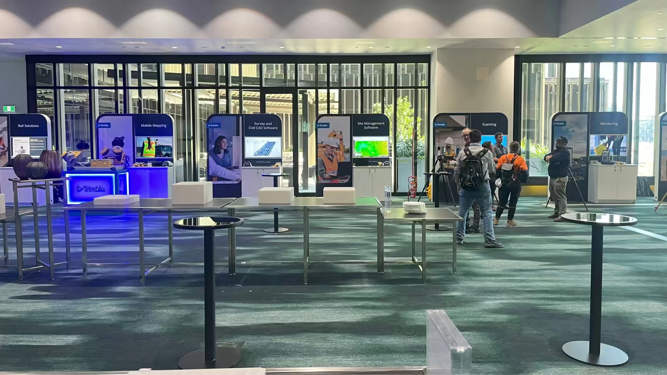An exhibition booth for Trimble in a modern hall featuring banners for Rail Solutions, Mobile Mapping, Scanning, and Monitoring with attendees.