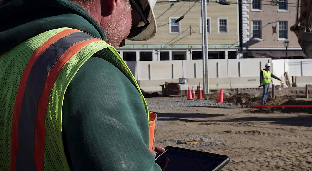 A construction worker in a high-visibility vest uses a Trimble tablet on a job site with orange cones and a yellow building in the background.