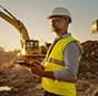 A construction professional wearing safety gear uses a Trimble rugged tablet on a job site with an excavator in the background.