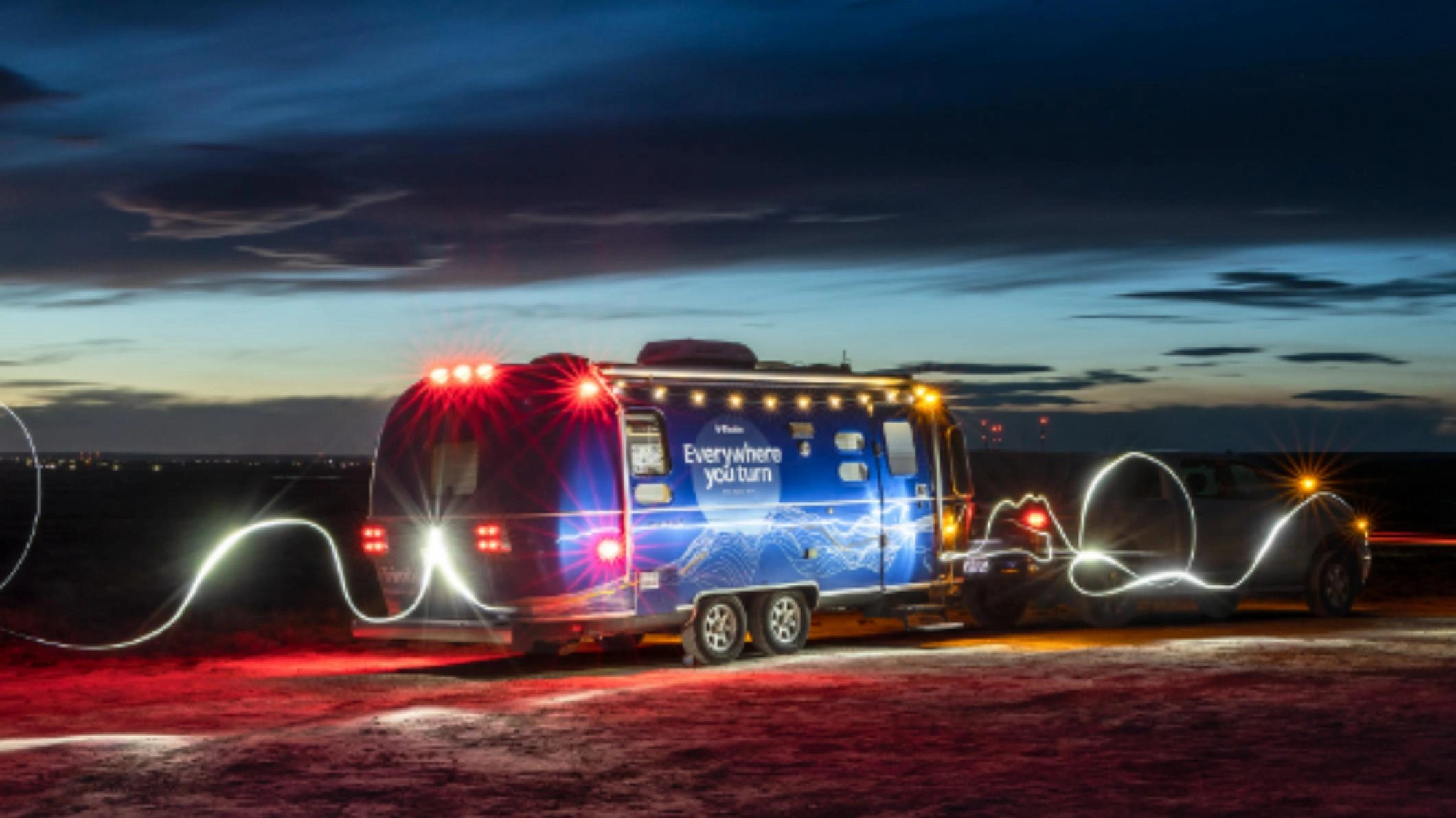 Ein grauer Lastwagen zieht in der Abenddämmerung den blauen Airstream-Trailer „Avery Weir“ von Trimble hinter sich her. Der Wagen ist mit dem Slogan „Everywhere You Turn“ versehen. Weiße Lichtspuren ergänzen das Bild.