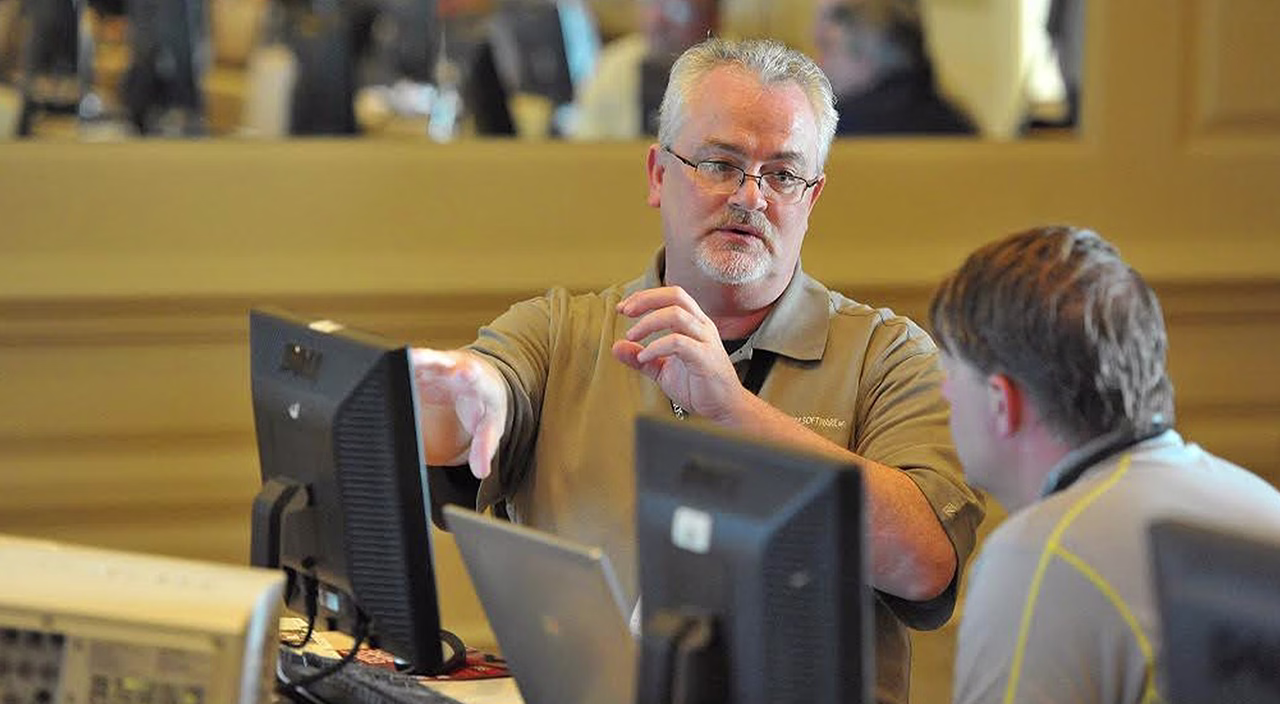 A software trainer wearing glasses and a tan polo shirt gestures toward a computer monitor while explaining a concept to a seated student.