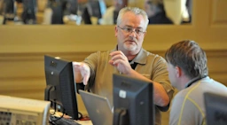 A software trainer wearing glasses and a tan polo shirt gestures toward a computer monitor while explaining a concept to a seated student.