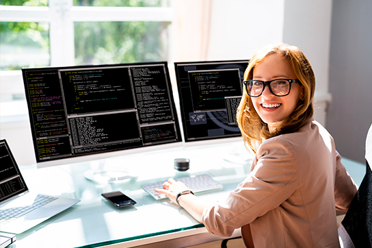 Smiling female software developer wearing glasses, looking over her shoulder while working at a desk with multiple monitors displaying code.