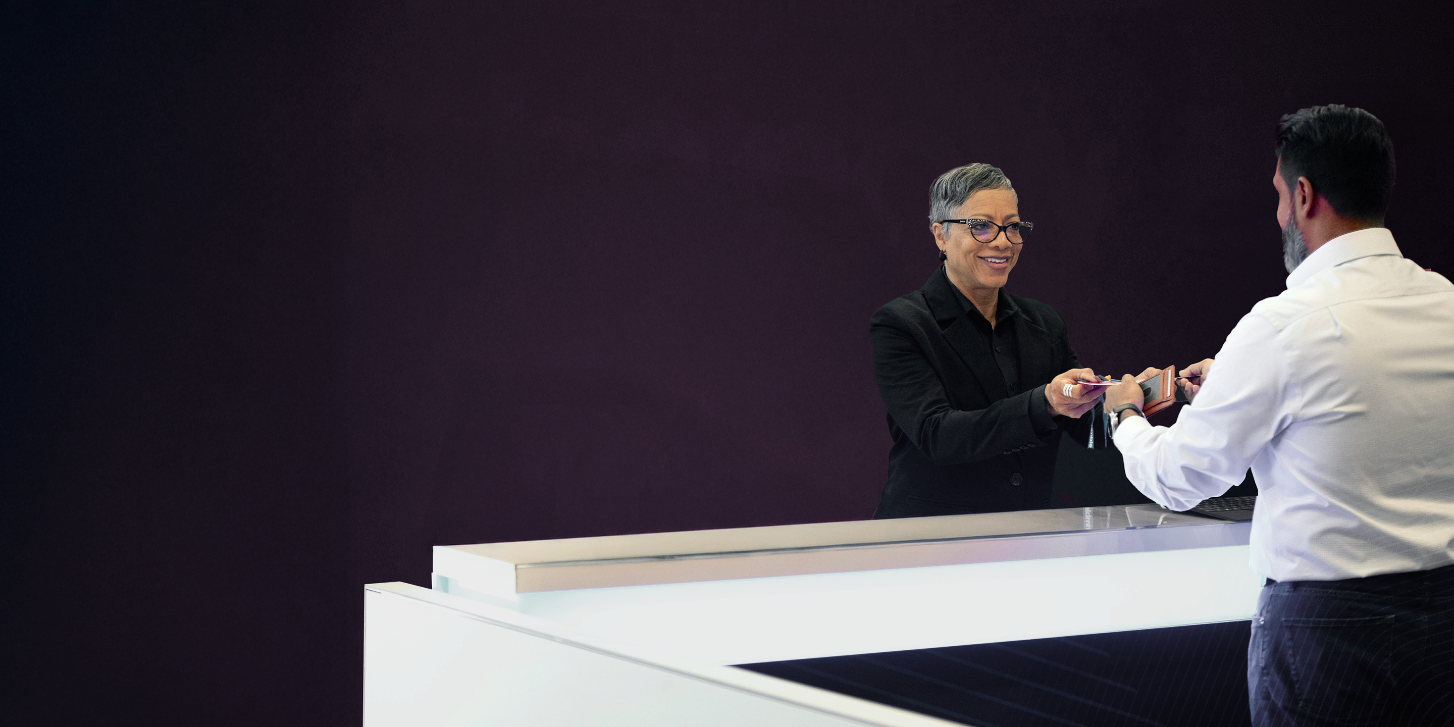 Smiling woman with short grey hair and glasses hands a card to a man at a modern Trimble reception desk with a dark purple background.