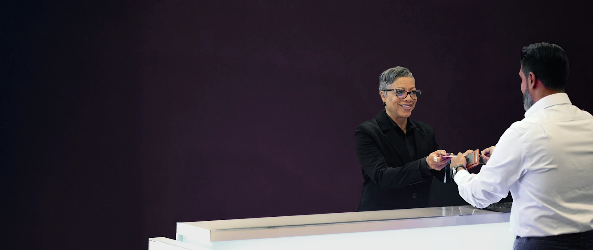 Smiling woman with short grey hair and glasses hands a card to a man at a modern Trimble reception desk with a dark purple background.