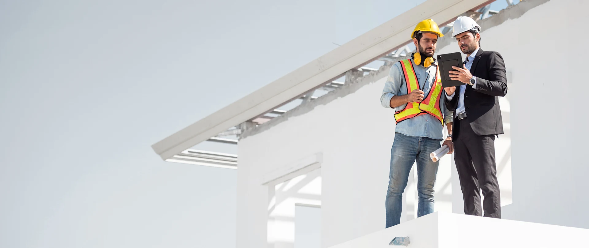 An engineer in a suit and a construction worker in a safety vest review plans on a tablet atop a white building under construction.