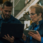 Two warehouse workers collaborate using a laptop and a tablet in an industrial environment.