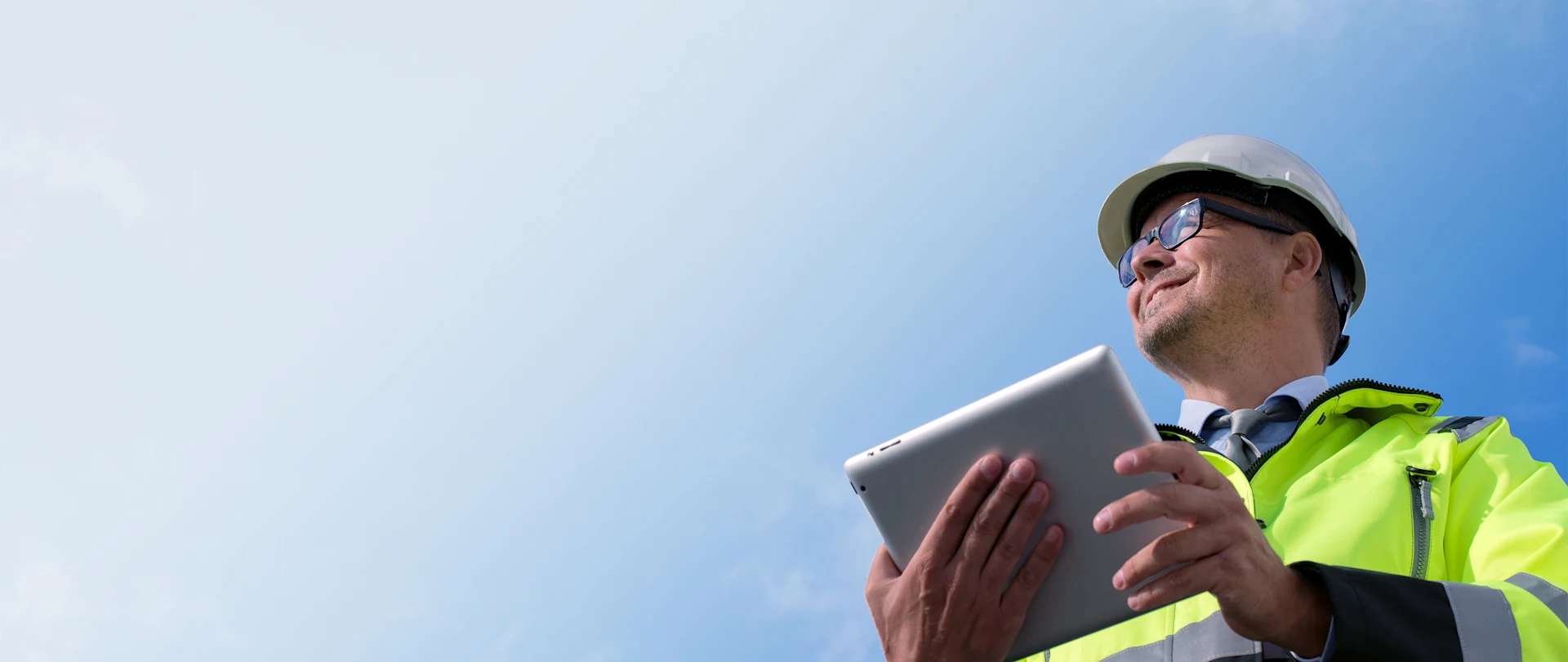 A construction professional in a white hard hat and neon vest uses a tablet for Trimble technology, set against a clear blue sky.