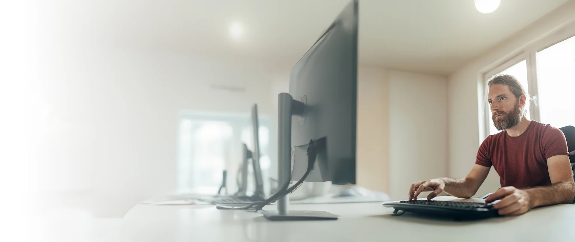 A man with a beard sitting at a desk and working on a desktop computer.