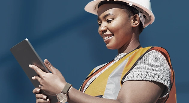 A smiling woman in a white hard hat and orange safety vest reviews information on a tablet against a solid blue background.