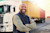 Smiling male truck driver with folded arms standing in front of his semi-truck and shipping containers at a depot.