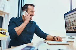 A smiling professional on a phone at a desk with an architectural model and a computer screen displaying Trimble's 3D Warehouse gallery.