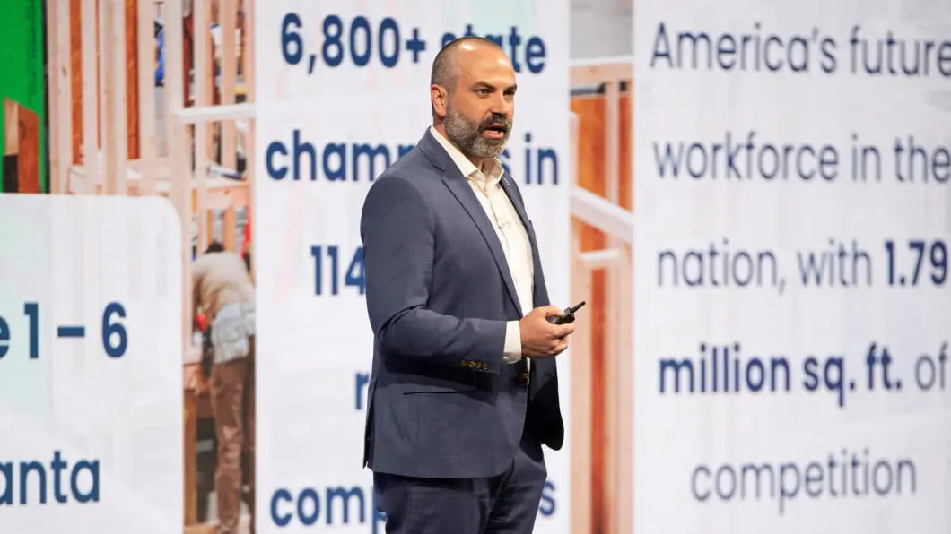A man presents at a SkillsUSA conference in Atlanta before a backdrop highlighting construction workforce statistics.