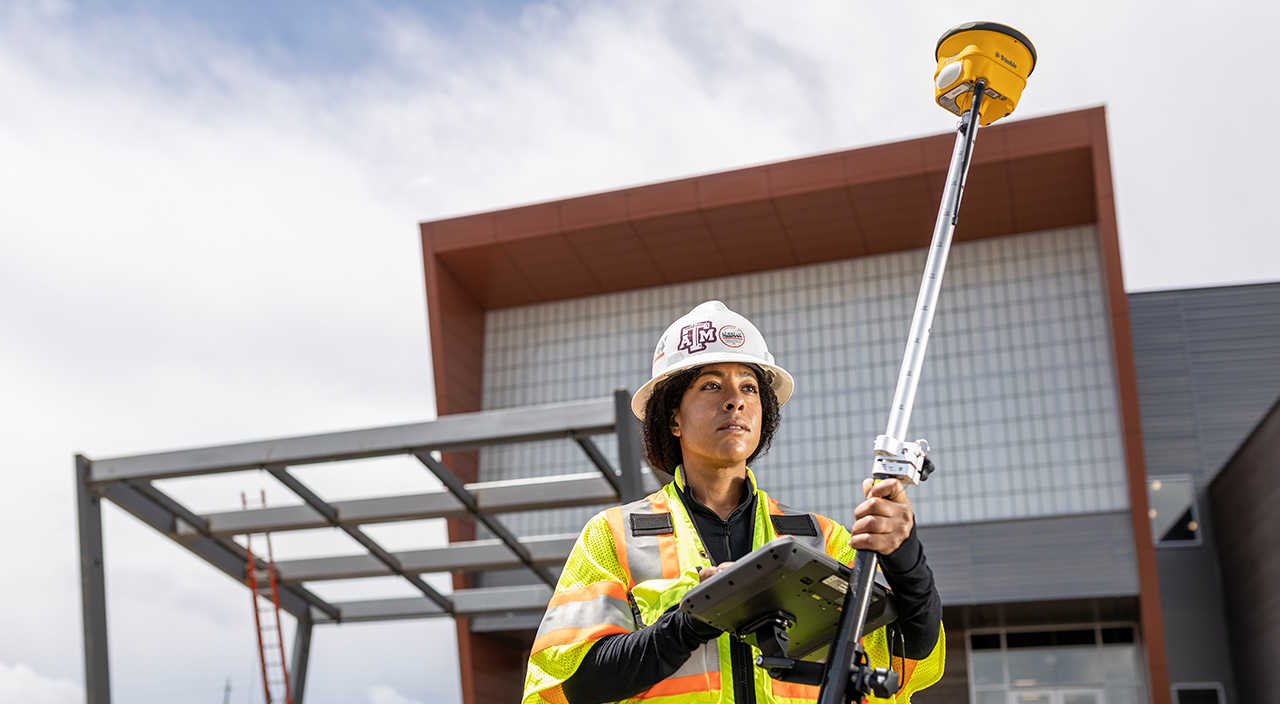 A woman in a Texas A&M hard hat uses a Trimble R12 GNSS receiver and TSC7 controller at a construction site.
