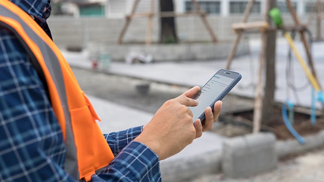A worker in an orange safety vest and plaid shirt uses a Trimble construction mobile app on a job site with staked trees in the background.