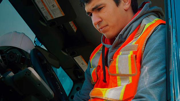 A close-up, eye-level shot shows a man in a gray hooded sweatshirt and a bright orange safety vest with reflective stripes, seated in the cab of a vehicle.
