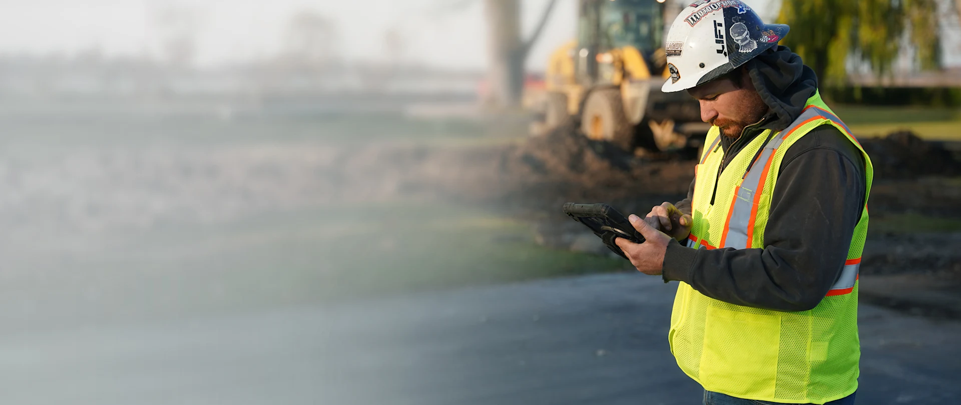 A construction worker uses a Trimble rugged tablet on-site, with a yellow wheel loader in the background.