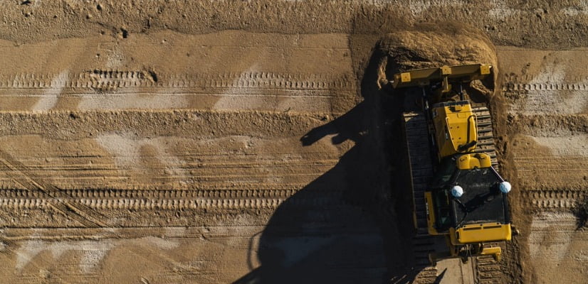 Aerial view of a yellow bulldozer pushing sand on a construction site, leaving track marks.