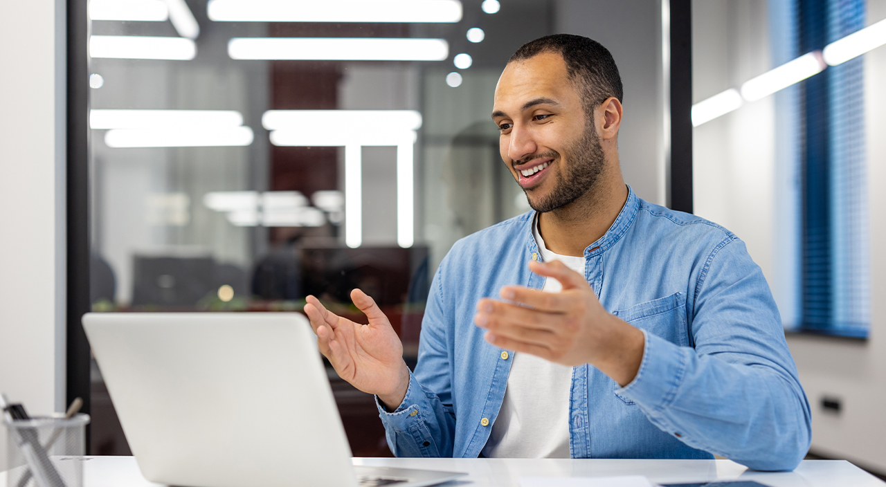 A smiling man in a blue denim shirt gestures during a video call on a laptop in a modern office setting with glass partitions.