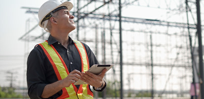 An engineer in a hard hat and safety vest looks up at an electrical substation while holding a tablet. (102 characters)