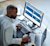 A man works at a desk using a monitor displaying the Trimble Construction One analytics dashboard, featuring project and equipment data.
