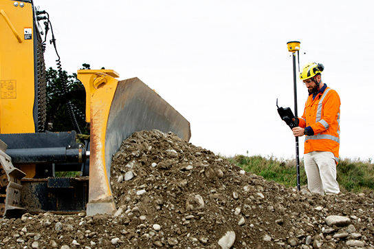 Construction worker in an orange safety vest operating surveying equipment near a bulldozer moving a pile of dirt and rocks