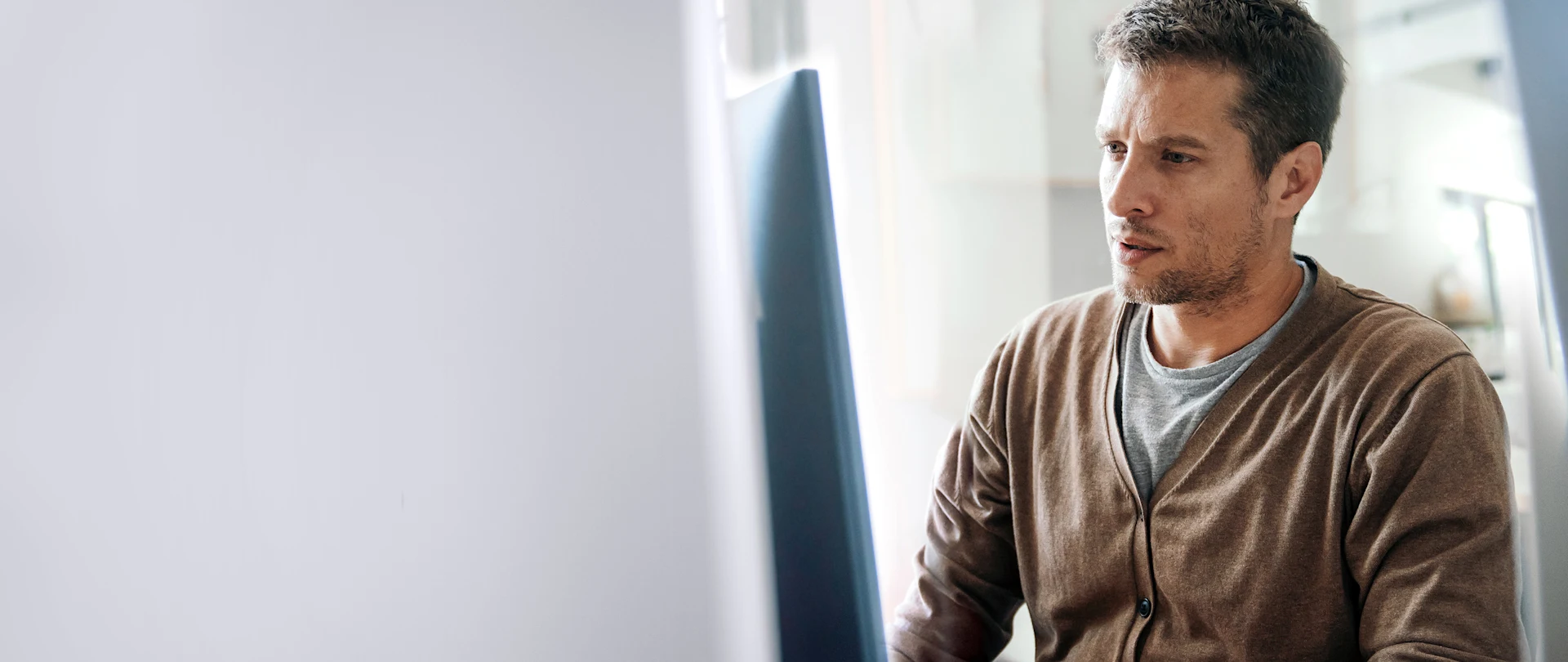 A man in a brown cardigan looks intently at a computer screen while working at a desk in a modern, brightly lit office.